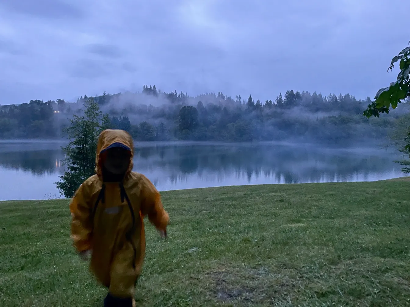 A child in a yellow rain suit running across a grassy field beside a misty lake at dusk, fog rolling through the evergreen trees