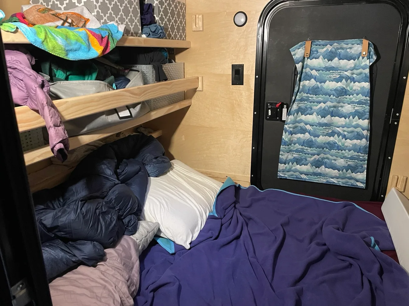 The main bed of a teardrop camper with a purple fleece blanket and a crumpled blue down quilt, storage shelves stacked with baskets and warm layers, and a mountain-print curtain hanging on the door