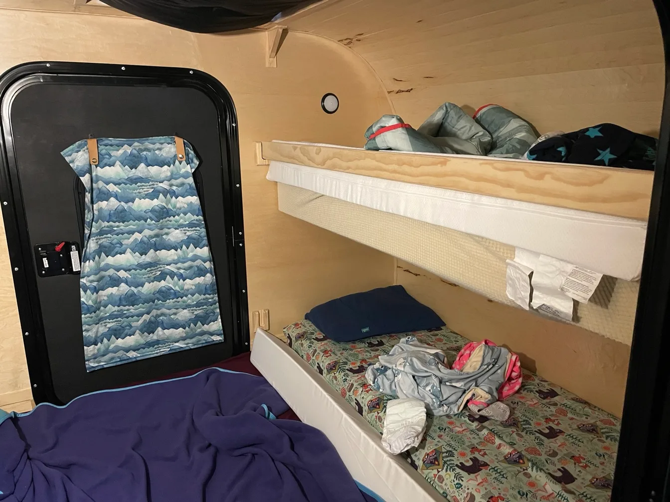 Inside a teardrop camper cabin, two kids' bunks along the wall with sleeping bags, a mountain-print cover on the door, and a purple fleece blanket on the main bed