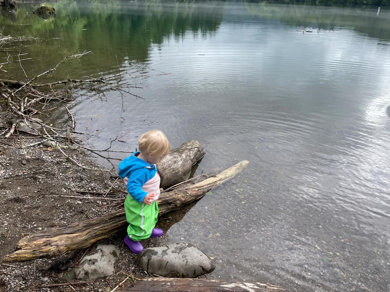 A toddler in a blue fleece and green rain pants leaning on a driftwood log at the edge of a calm lake, purple rain boots on
