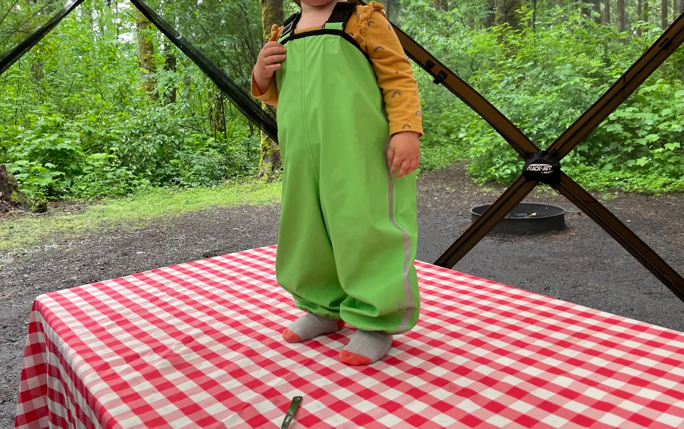 A toddler standing on a gingham-covered camp table wearing bright green rain overalls with reflective strips over a long-sleeve shirt